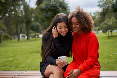 Smiling friends using phone sitting on bench at park