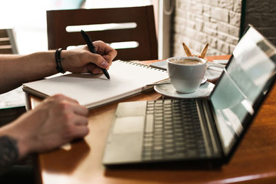 Unrecognizable man sketching in notepad near cup of coffee and laptop while sitting at table an working on remote project in cafeteria