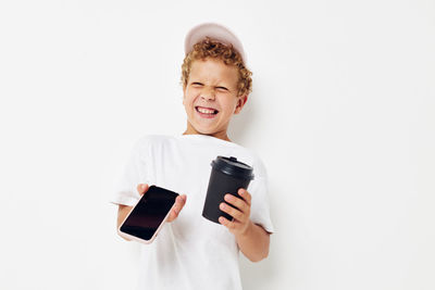 Cheerful boy holding mobile phone with coffee cup against white background