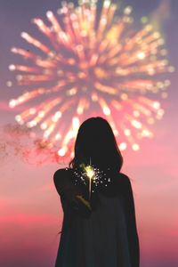 Woman holding illuminated sparkler against dramatic sky at sunset