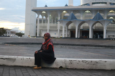 Portrait of young woman sitting at roadside against mosque in city