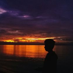 Silhouette boy standing in sea against sky during sunset