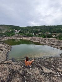 High angle view of woman sitting on rock by pond against sky