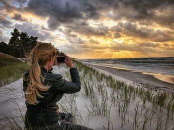 Man photographing at beach against sky during sunset