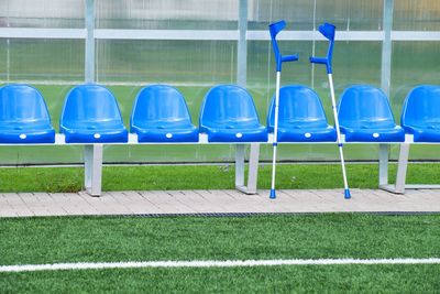 Forearm crutch at blue plastic seats on outdoor stadium players bench, chairs below transparent roof