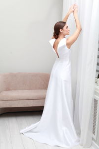 Full length portrait of bride in elegant wedding dress posing with arms raised, pensive expression 