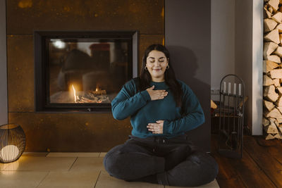 Woman with eyes closed doing breathing exercise while sitting cross-legged near fireplace at home