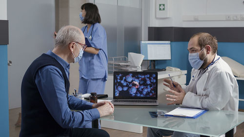 Male doctor discussing over laptop with patient in medical clinic during pandemic