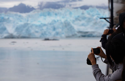 Rear view of people photographing glacier while standing on boat