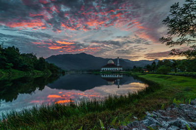 Mosque by river against cloudy sky during sunrise