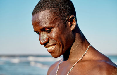 Happy african american man with silver chain on neck smiling on blurred background of sea and cloudless blue sky in summer