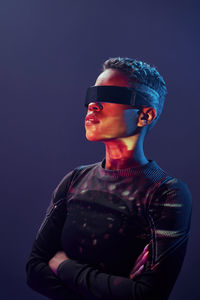 Serious african american female with short hair wearing contemporary vr glasses while standing with crossed arms on purple background in studio with glowing neon lights