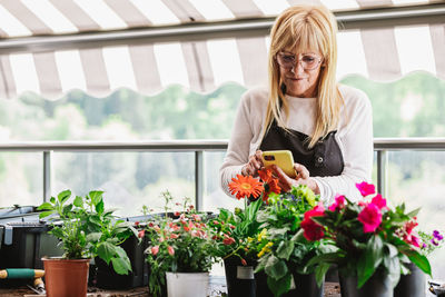 Busy female gardener with blond hair in apron browsing phone while standing near various potted flowers in light room with window