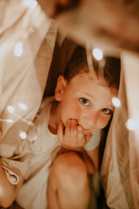 Cute preschool boy in white t-shirt inside shelter made of crumpled sheets with garlands and lights