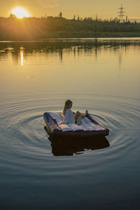 Rear view of young woman sits on a mattress on the lake during sunset