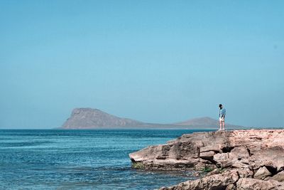 Rear view of woman standing on rock by sea against clear sky