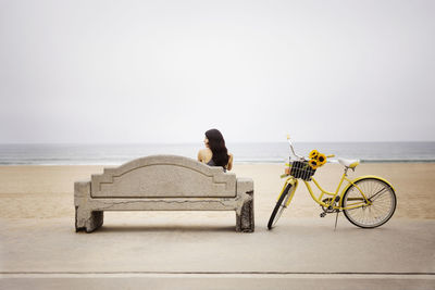 Rear view of woman sitting on bench at beach against clear sky