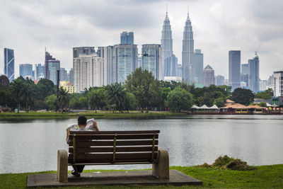 Rear view of man reading newspaper while sitting on bench by lake in city