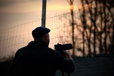 Rear view of man photographing while standing by fence against sky during sunset