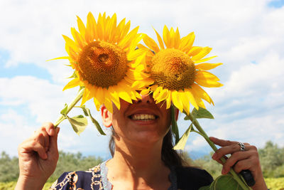 Close-up portrait of smiling woman holding sunflower against sky