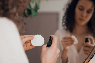 Young woman doing self-care routine at mirror, using aroma oil or serum for skin