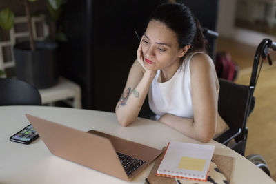 Woman with hand on chin watching laptop while sitting on wheelchair at home