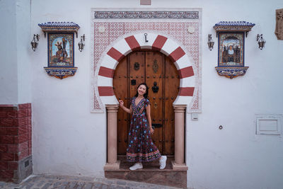 Full body of smiling asian female traveler looking at camera while standing near old ornamental building during trip in spain