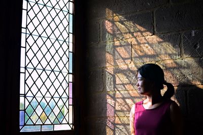 Young woman looking through window while standing against wall