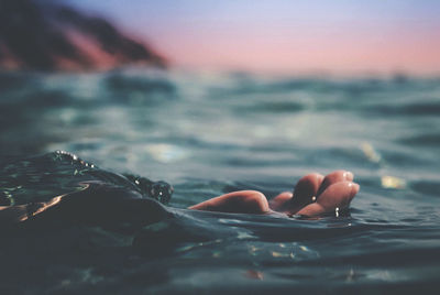 Cropped hand in sea against sky during sunset