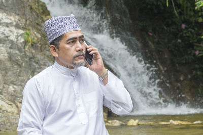 Thoughtful man talking on phone while sitting against waterfall