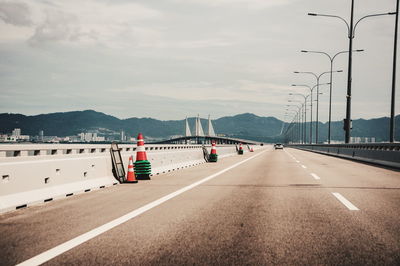 Traffic cones on bridge by street lights leading towards mountains against sky