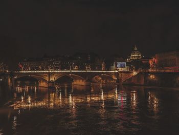 Illuminated bridge over river by buildings against sky at night