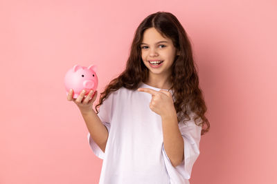 Portrait of smiling young woman holding piggy bank against pink background