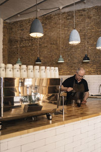 Concentrated mature male barista in apron pouring hot aromatic coffee from coffee maker into glass placed on wooden counter in cafe