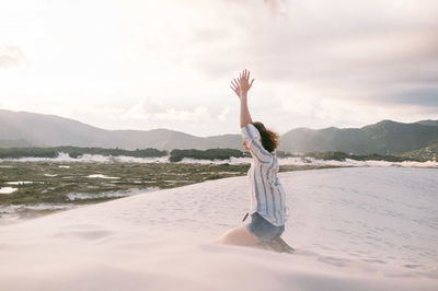 Side view of woman with arms raised kneeling on sand against cloudy sky