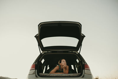Thoughtful woman looking away while lying in car trunk against clear sky