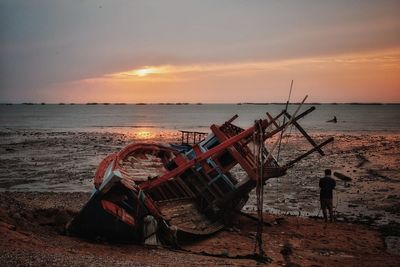 Man standing by shipwreck at beach against sky during sunset