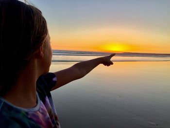 Girl pointing while standing at beach against sky during sunset