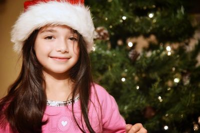 Portrait of smiling girl wearing santa hat against illuminated christmas tree at home