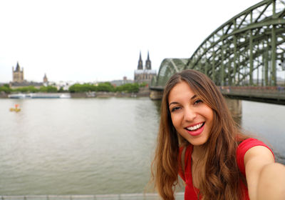 Portrait of smiling young woman against bridge over river in city