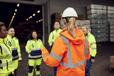 Rear view of female engineer in reflective clothing discussing with workers at industry