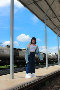 Young woman wearing sunglasses standing at railroad station against sky