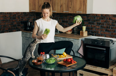 Cheerful young woman preparing green salad at home and jokingly throwing vegetables up