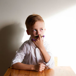 Thoughtful boy wearing flowers mustache while sitting at desk against wall