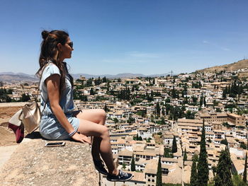 Side view of young woman sitting on retaining wall against sky in city