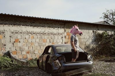 Man wearing horse mask standing in abandoned car against sky