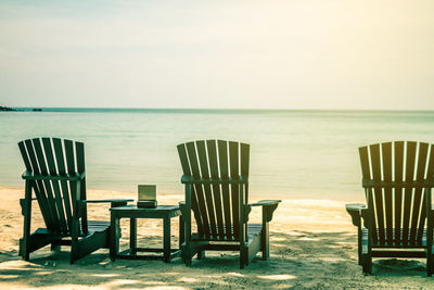 Empty adirondack chairs on beach against sky