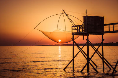 Silhouette fishing net over sea against orange clear sky during sunset