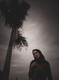 Portrait of young man standing by palm tree against sky