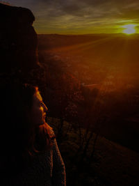 Thoughtful woman looking away while sitting on mountain against sky during sunset